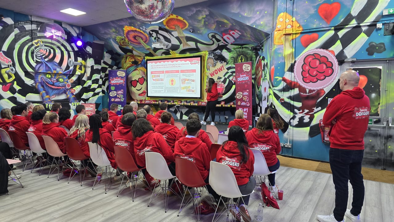 A team in red hoodies seated in rows facing the presentation screen in the hall, the murals either side